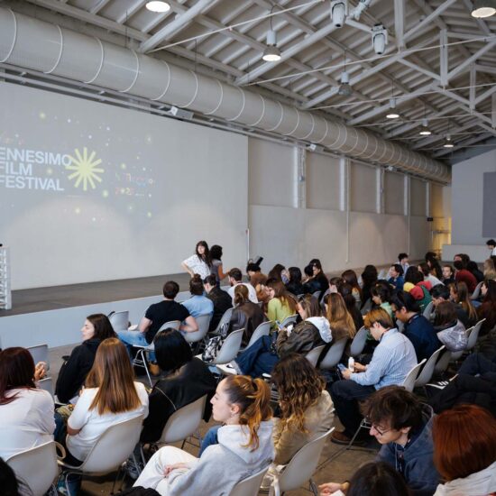 Pubblico numeroso seduto in sala durante il "Ennesimo Film Festival", con un palco e un proiettore sullo sfondo.