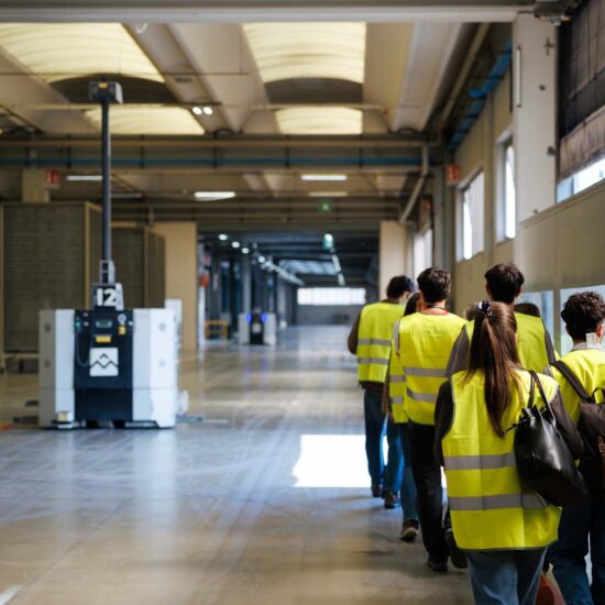 Gruppo di persone in gilet riflettenti che cammina in un ampio ambiente industriale con un robot autonomo in primo piano.