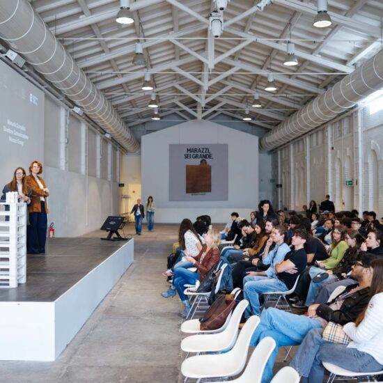 Tre relatrici sul palco durante un evento, mentre il pubblico ascolta attentamente in una sala luminosa.