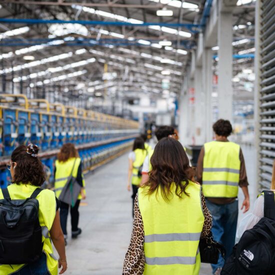 Gruppo di persone in giubbotti gialli che camminano in un'industria, con macchinari blu visibili in background.