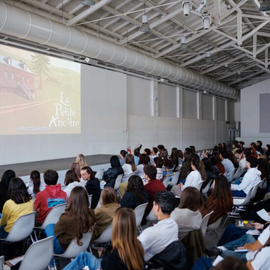 Pubblico in sala durante una presentazione di "La Petite Ancêtre", con un'immagine proiettata su schermo.