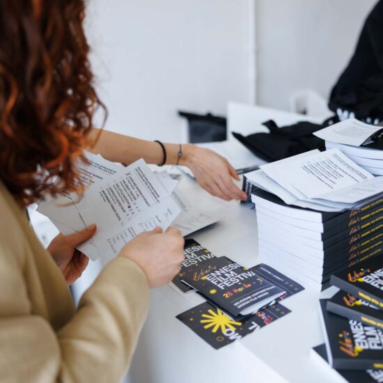 Una persona con capelli ricci conta dei documenti accanto a un tavolo con libri e materiali promozionali di un festival.